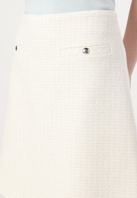 Jupe blanche texturée en matériau tweed, avec deux fausses poches et des accents de boutons en argent à l'avant.