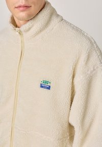 Svetlokrémová fleecová bunda s vysokým golierom, zapínaním na zips a malou modro-zelenou nášivkou na hrudi. Mäkká, textúrovaná tkanina.