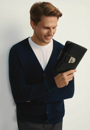 Granatowy kardigan na białej koszuli, trzymający czarny skórzany portfel z detalem z srebrnej klamry. Gładka tekstura, prostokątny kształt.