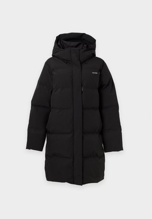 Femme portant un long manteau matelassé noir avec capuche, un pull gris, un pantalon noir et des chaussures noires, debout devant un fond blanc uni.