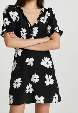 Femme portant une robe noire à grands imprimés floraux blancs, manches courtes bouffantes et col en V à volants, bras croisés.