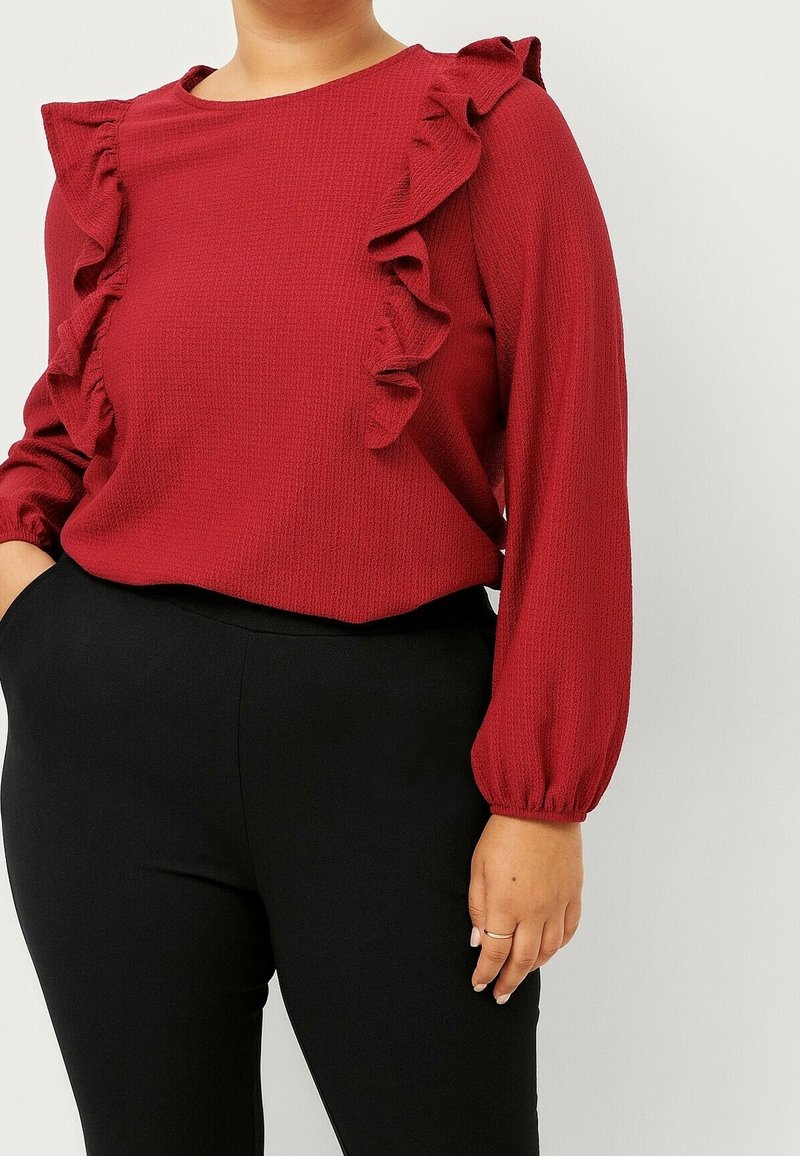 Personne vêtue d'un chemisier rouge texturé à manches longues avec des détails froncés sur le devant et un pantalon noir ajusté, debout devant un fond uni.