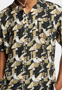 Camicia a maniche corte con motivo geometrico di case in tonalità terrose di beige, oliva e nero. Tessuto morbido con taschino sul petto.
