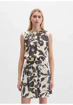 Jeune femme portant une robe crème sans manches avec un grand imprimé floral noir, un col froncé et une taille élastique, debout devant un fond uni.