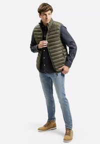 Oliwkowa, pikowana kamizelka na ciemnej koszuli z guzikami, zestawiona z jasnoniebieskimi dżinsami i brązowymi butami. Kamizelka ma poziome pikowane segmenty.