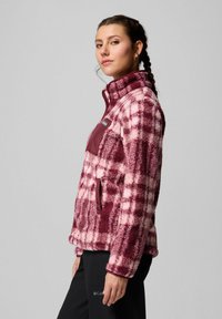 Donna con i capelli intrecciati che indossa una giacca di pile a scacchi bordeaux e rosa e pantaloni neri, voltata di lato su uno sfondo semplice.