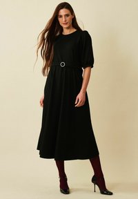 Musta midi-mekko puff-hihnoilla ja napakalla vyötäröllä, jossa on pyöreä vyönsolki. Yhdistetty burgundin värisiin nyloneihin ja mustiin korkokenkiin.