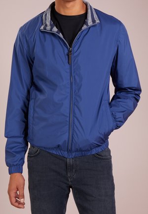 Homme portant une veste zippée bleue avec des poignets élastiques et un col rayé, chemise noire en dessous et jean foncé.
