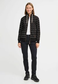 Giacca nera con zip, decorata con un motivo a zigzag, maniche elastiche e cappuccio con cordino. Combinata con pantaloni scuri e scarpe nere. Texture liscia.
