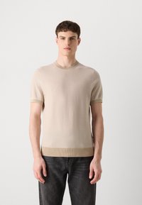 Beige kötött rövid ujjú pulóver kerek nyakkivágással, textúrázott felülettel és kontrasztos bordázott mandzsettákkal és hem. Sötét farmernadrággal viselve.
