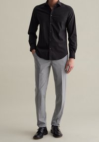 Schwarzes, langärmliges Hemd mit Knöpfen und einem Standardkragen, kombiniert mit grauen Nadelstreifenhosen und schwarzen Schuhen, vor einem neutralen Hintergrund stehend.