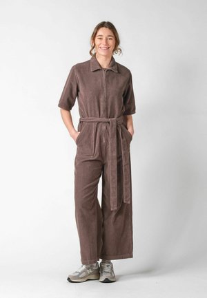 Žena stojaca s rukami v kapsách, oblečená v hnedom kordurovom overal s opaskom a sivými teniskami, usmievajúca sa na bielom pozadí.