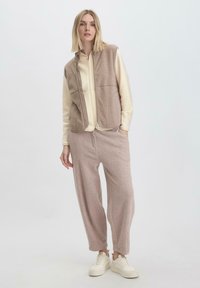 Beige Fleece-Weste mit Reißverschluss über einem cremefarbenen Langarmoberteil, kombiniert mit hellbeigen elastischen Hosen und weißen Sneakers.