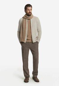 Beżowy wełniany sweter rozpinany na guziki noszony na brązowym golfie, do tego brązowe spodnie i pasujący szalik oraz proste brązowe buty.