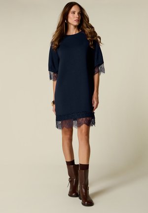 Woman with long hair wearing a navy blue dress with lace trim on sleeves and hem, paired with brown ankle boots and dark socks.