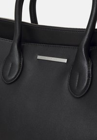 Černá kožená taška s hladkým povrchem, dvěma uchy a kovovým štítkem s názvem značky. Zipové uzavření nahoře.