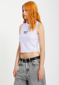 Wit cropped tanktop met een zwart logo op de borst, gecombineerd met high-waisted grijze jeans en een zwarte riem.