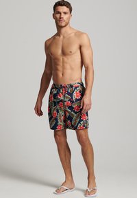 Costume da bagno da uomo con uno sfondo blu navy scuro, caratterizzato da grandi fiori rossi e motivi di foglie verdi. Realizzati in tessuto leggero con vita regolabile tramite coulisse.
