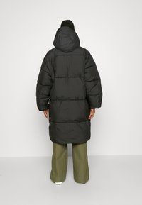 Zwarte pufferjas met een capuchon, met een lang ontwerp en brede mouwen. Het materiaal lijkt glad met een gewatteerd patroon.