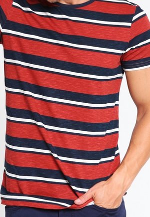 Homme portant un t-shirt à manches courtes avec des rayures horizontales rouges, bleu marine et blanches, une main dans la poche, sur un fond blanc.