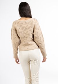 Pulover tricotat bej cu un model texturat în valuri, având un decolteu larg și mâneci tip balon, purtat cu pantaloni haioși de culoare deschisă.