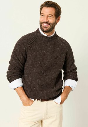 Mann mit Bart lächelnd, trägt einen dunkelbraunen gestrickten Pullover über einem weißen Hemd, mit Händen in den Taschen einer beigen Hose vor schlichtem Hintergrund.