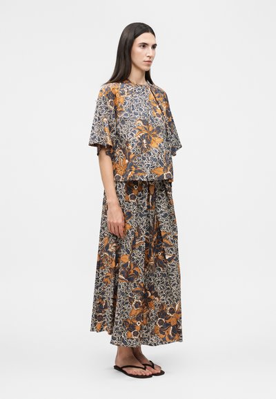 Γυναίκα με μακριά σκούρα μαλλιά που φοράει ένα χαλαρό τοπ και μακριά φούστα με εναρμονισμένο λουλουδάτο μοτίβο σε αποχρώσεις πορτοκαλί, ναυτικό και κρεμ, όρθια πλαγίως.