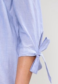 Hellblaue Leinenbluse mit hochgekrempelten Ärmeln, die ein Bindeband am Bündchen aufweist. Der Stoff hat ein weiches, strukturiertes Aussehen.