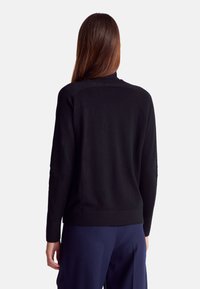 Černý pletený svetr s vysokým výstřihem, zdobený texturovanými detaily a žebrovaným lemem. Modelka je nafocena zezadu, se spárovanými námořnickými kalhotami.