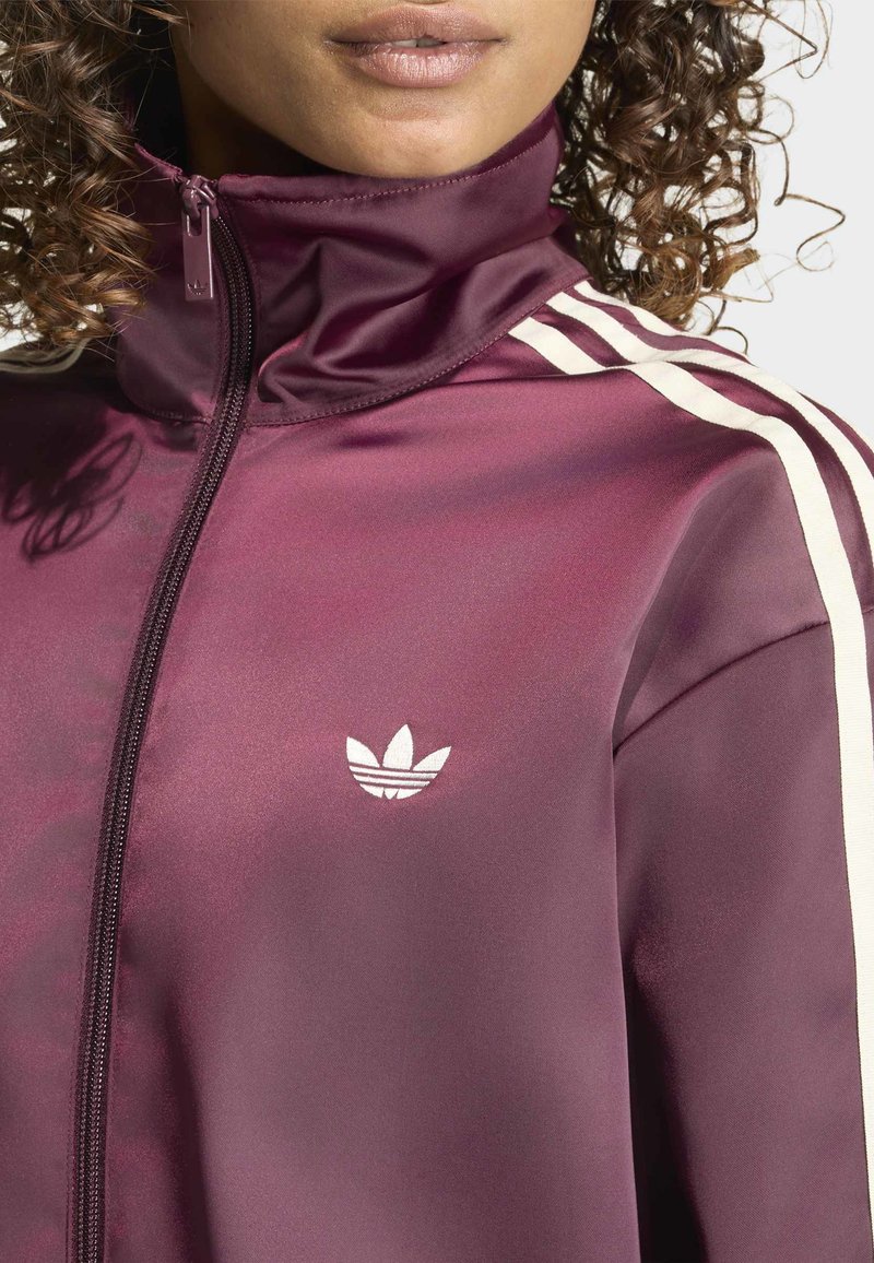 Gros plan d'une personne portant une veste de survêtement bordeaux avec des bandes blanches sur les manches et un logo Adidas blanc sur la poitrine.
