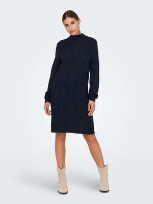 Femme portant une robe-pull à manches longues en maille torsadée bleu marine, associée à des bottines beige, debout avec une jambe légèrement en avant.