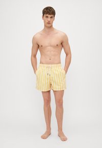 Gelb-weiße gestreifte Badehose mit elastischem Bund, Kordelzug und Seitentaschen. Leichte Stoffqualität und lockere Passform.