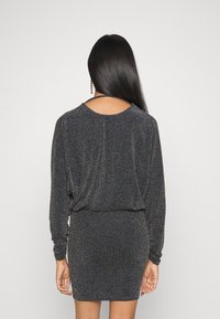 Svart metallisk klänning med långa ärmar, samlad midja och öppen ryggdetalj, som visar upp ett glänsande texturerat tyg.