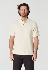 Krämfärgad polo-tröja med korta ärmar, strukturerad tyg, tre-knappsknäppning med svarta knappar och en liten märkestag i nederkant.