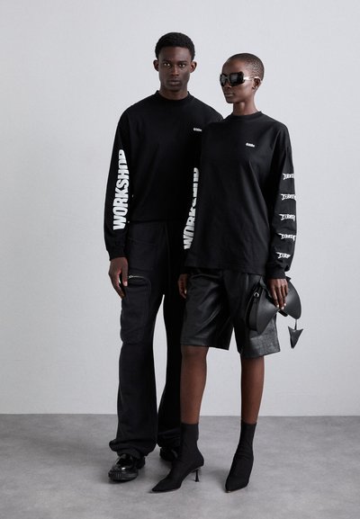 Black long-sleeve shirts feature contrasting white graphics. One model wears black cargo pants; the other wears black leather shorts and heeled boots.