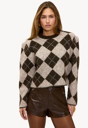 Vrouw draagt een bruine en beige argyle gebreide trui met geplooide schouders en donkerbruine leren shorts, staand tegen een effen achtergrond.