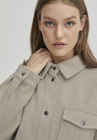 Beige tekstureeritud jakk kraega, mustade nööpide ja rinnataskuga. Valmistatud pehmest ja soojast kangast, edastades mugava välimuse.