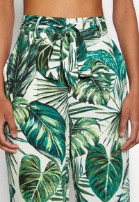 Pantalon taille haute à jambes larges arborant un motif de feuilles vertes sur fond blanc, avec une ceinture nouée à l'avant et des plis pour ajouter de la texture.