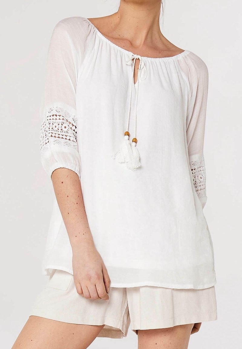 Blouse blanche à coupe ample, col froncé et détails en crochet sur les manches. Dotée d'un lien avec des pompons et d'un tissu léger.