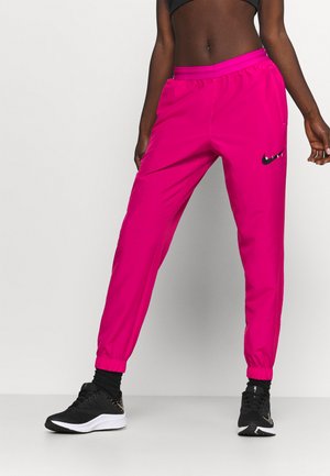 Persoon draagt felroze Nike joggingbroek, zwarte sokken en zwarte Nike sneakers, staand met één been iets naar voren tegen een witte achtergrond.