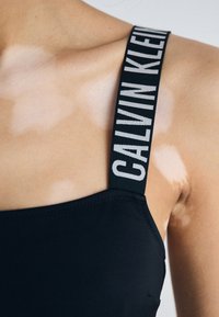 Schwarzer Badeanzug mit einem breiten schwarzen Träger, auf dem "CALVIN KLEIN" in weißen Buchstaben steht. Das Material wirkt glatt mit einem figurbetonten Design.