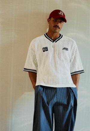 Uomo con baffi che indossa un berretto rosso, una maglia bianca a maniche corte con il "03" sul petto e pantaloni larghi blu a righe, in piedi contro un muro beige.