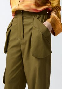 Maslinasto zeleni hlače s velikim, oštrim džepovima, zatvaračem sprijeda i opuštenim krojem. Tkanina izgleda glatko i lagano.