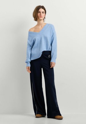Vrouw draagt een lichtblauwe off-shoulder trui, zwarte wijde broek met witte streep en bruine schoenen, staand tegen een effen muur.