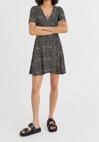 Schwarzes Blumen Kleid mit V-Ausschnitt, kurzen Ärmeln und einem ausgestellten Rock. Kombiniert mit schwarzen Riemensandalen, die zwei breite Riemen haben.