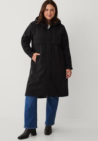 Sort vandtæt lang frakk med hætte, frontlommer og lynlåsdetaljer. Stylet over blå jeans og brune ankelstøvler med hæl.