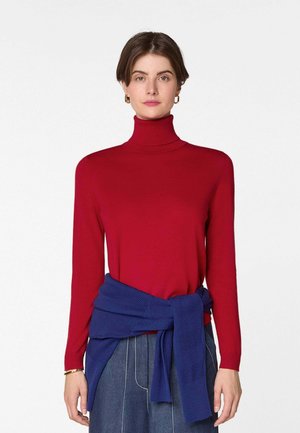 Vrouw draagt een rode coltrui en een blauwe gebreide trui om haar middel geknoopt, staande tegen een effen witte achtergrond.