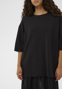 Schwarzes übergroßes T-Shirt aus weichem Stoff mit rundem Ausschnitt und ellbogenlangen Ärmeln, getragen über einem transparenten, schwarzen, mehrlagigen Rock.