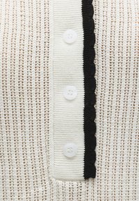 Κοντινή λήψη λευκού πλεκτού υφάσματος με κάθετη λωρίδα με τρία λευκά κουμπιά και μαύρο διακοσμητικό κυματιστό τελείωμα.