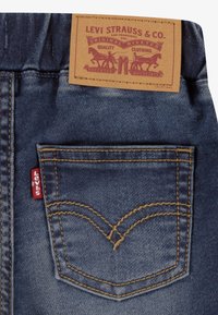 Jean en denim avec une pièce en cuir marron embossée du logo, des coutures dorées, et une étiquette rouge sur la poche arrière avec un motif de couture incurvée.